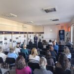 Sala audytoryjna pełna ludzi. Przodem do nich stoi mężczyzna.