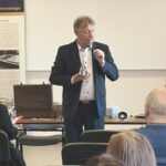 Mężczyzna w garniturze stoi w sali przed publicznością.