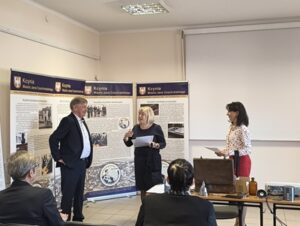 Blondwłosa kobieta wręcza dokument mężczyźnie w garniturze. Obok drugi stoi mężczyzna.