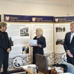 Blondwłosa kobieta wręcza dokument mężczyźnie w garniturze. Obok drugi stoi mężczyzna.