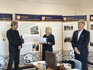 Blondwłosa kobieta wręcza dokument mężczyźnie w garniturze. Obok drugi stoi mężczyzna.