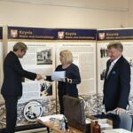 Blondwłosa kobieta wręcza dokument mężczyźnie w garniturze. Obok drugi stoi mężczyzna.