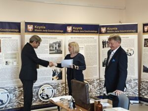 Blondwłosa kobieta wręcza dokument mężczyźnie w garniturze. Obok drugi stoi mężczyzna.