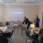 Dwie kobiety stoją na przedzie sali, jedna pochyla się nad laptopem. W tle siedzą dzieci w grupach.