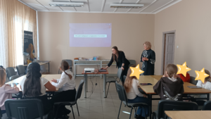 Dwie kobiety stoją na przedzie sali, jedna pochyla się nad laptopem. W tle siedzą dzieci w grupach.