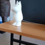 Figurka muminka stoi na stoliku przed laptopem. W tle kobieta w brązowej sukience.