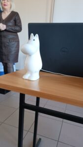 Figurka muminka stoi na stoliku przed laptopem. W tle kobieta w brązowej sukience.