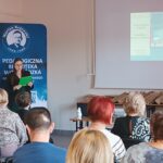 Kobieta w zielonej sukience stoi przed widownią i czyta z zielonej podkładki.