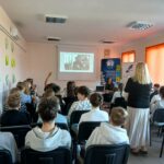 Blondwłosa kobieta stoi, wokół siedzi młodzież na krzesłach.