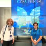 Dwie kobiety pozują przed ekranem wyświetlającym plakat kampanii "Cała Polska czyta dzieciom".