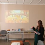 Kobieta w zielonej sukience odczytuje informacje ze slajdu trzymając książki.