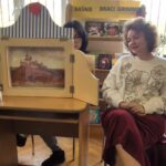 Kobieta w białym sweterku siedzi przy teatrzyku kamishibai.