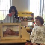 Kobieta siedzi przy teatrzyku kamishibai - druga stoi za nim.