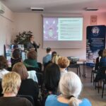 Kobieta w garniturze i okularach prezentuje wykład przed pełną salą audytoryjną.