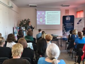 Kobieta w garniturze i okularach prezentuje wykład przed pełną salą audytoryjną.