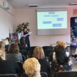 Kobieta w różowym swetrze, czyta z kartki stojąc przed siedzącymi ludźmi.