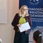 Blondynka w okularach trzyma kartki, stojąc przed niebieskim banerem.