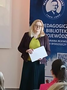 Blondynka w okularach trzyma kartki, stojąc przed niebieskim banerem.