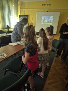 Nad stołem z materiałami plastycznymi pochyla się kobieta i trzy dziewczynki.