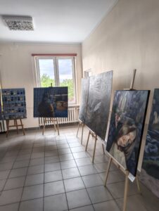 Okno w tle, na pierwszym planie obrazy na sztalugach.