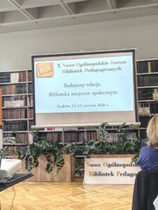 Ekran z wyświetloną prezentacją. W tle regały z książkami i kwiaty.