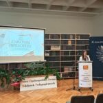 Kobieta w białej marynarce, stoi za katedrą i wygłasza referat.