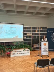 Kobieta w białej marynarce, stoi za katedrą i wygłasza referat.