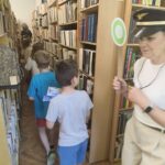Kobieta ubrana w czapkę konduktora zamyka pochód dzieci wśród regałów z książkami.