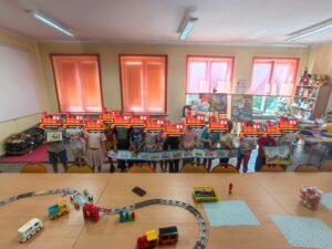 Grupa dzieci pozuje do zdjęcia, trzymając w rękach niebieski brystol z kolorowymi wagonami.