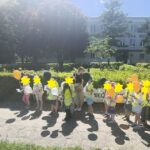 Grupa dzieci w kamizelkach odblaskowych i z balonami stoi na tle żywopłotu.