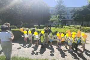 Grupa dzieci w kamizelkach odblaskowych i z balonami stoi na tle żywopłotu.