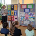 Blondwłosa kobieta stoi przy tablicach z pracami plastycznymi. Jest zwrócona twarzą do siedzących dzieci.