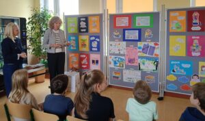 Blondwłosa kobieta stoi przy tablicach z pracami plastycznymi. Jest zwrócona twarzą do siedzących dzieci.