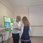 Kobieta w białej bluzce i kobieta w brązowej bluzce wręczają nagrodę dziewczynce w granatowej sukience.