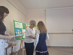 Kobieta w białej bluzce i kobieta w brązowej bluzce wręczają nagrodę dziewczynce w granatowej sukience.