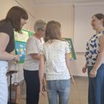 Trzy kobiety (w brązowej, białej i biało-niebieskiej koszuli) stoją wokół dziewczynki w białej koszulce.