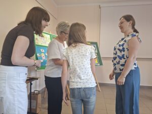 Trzy kobiety (w brązowej, białej i biało-niebieskiej koszuli) stoją wokół dziewczynki w białej koszulce.