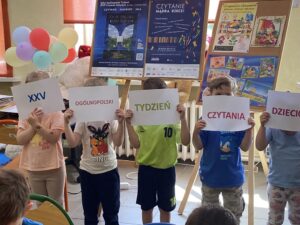 Pięcioro dzieci zasłania sobie twarze kartkami z hasłem "XXV Ogólnopolski Tydzień Czytania Dzieciom".