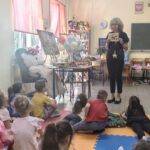 Stojąca kobieta pokazuje siedzącym dzieciom książkę.
