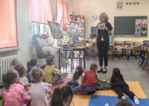 Stojąca kobieta pokazuje siedzącym dzieciom książkę.