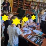 Grupa dzieci stoi wokół biurka i podsuwa kartki do opieczętowania. Nad nimi stoją 3 kobiety.