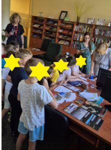 Grupa dzieci stoi wokół biurka i podsuwa kartki do opieczętowania. Nad nimi stoją 3 kobiety.