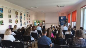 Kobieta w białej marynarce stoi przed siedzącymi ludźmi.