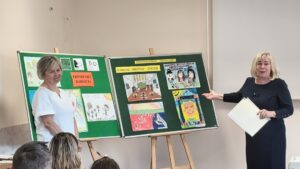 Dwie kobiety pokazują na zielone tablice wypełnione pracami plastycznymi.