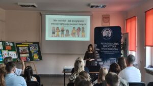 Sala audytoryjna, ludzie siedzą na krzesłach, na froncie stoi kobieta w ciemnej sukience.