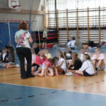 Kobieta w białej bluzce stoi nad grupą dzieci i im coś pokazuje.