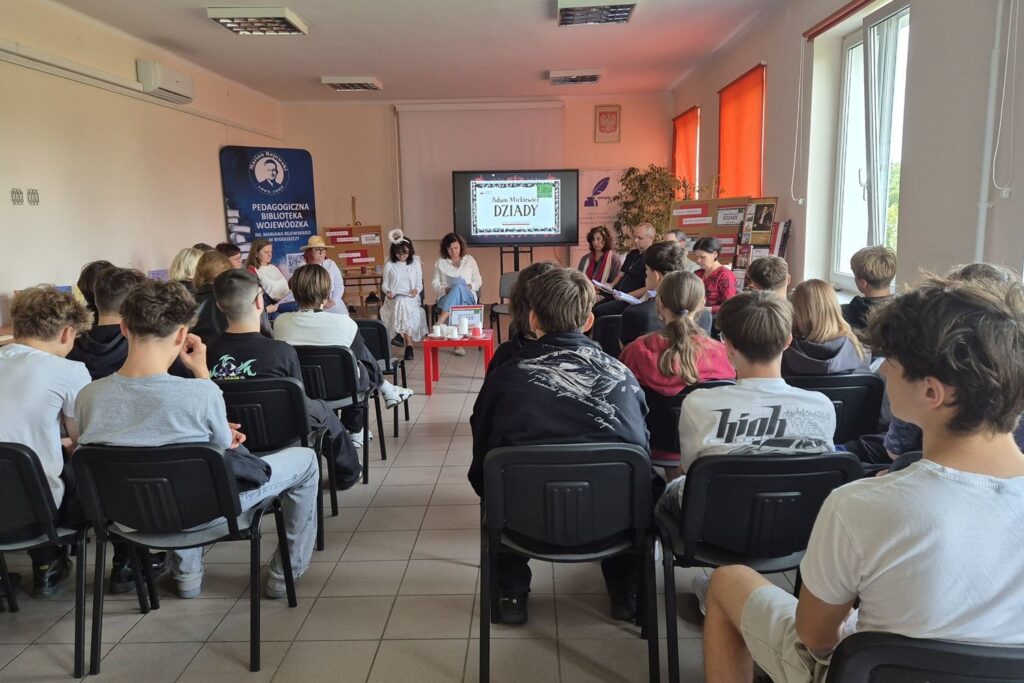 W sali młodzież obserwuje dorosłych siedzących w półokręgu. Na ekranie logo Narodowego Czytania 2026 "Dziady".