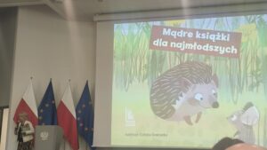 Kobieta na tle flag, mówi do mikrofonu i pokazuje prezentację multimedialną.