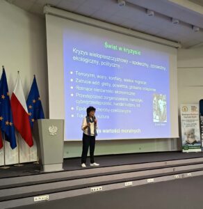 Kobieta w białym swetrze na schodach, za nią niebieski ekran.