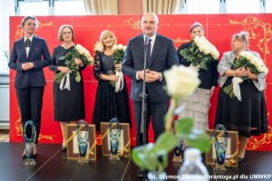 Mężczyzna w garniturze mówi do mikrofonu, z anim kilka kobiet.